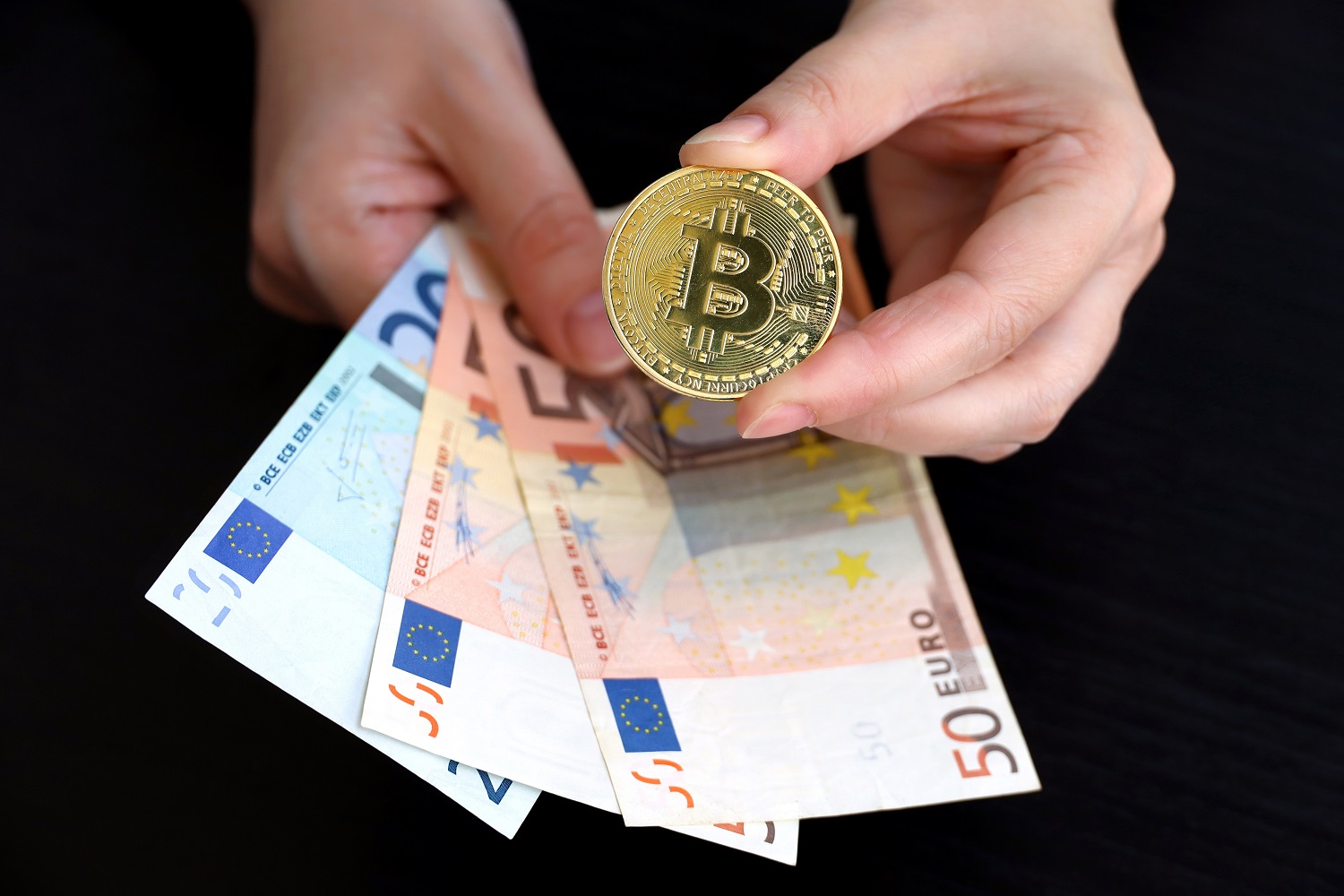 A person&rsquo;s hands holding a token intended to represent Bitcoin in one hand and euro banknotes in the other.